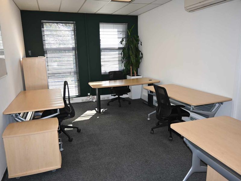 A functional office space featuring carpeted flooring, suspended ceiling, air conditioning, and large windows with venetian blinds.