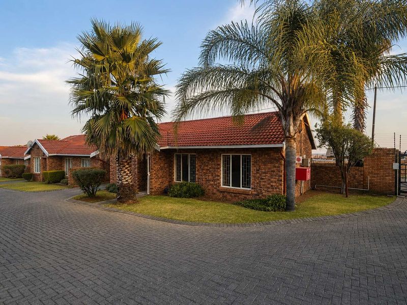 Front view of a face brick townhouse complex featuring a paved driveway, tiled roof, and secure perimeter fencing with a gated entrance.