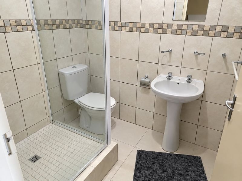 A functional bathroom featuring a pedestal basin, a glass-enclosed shower, and neutral wall tiling with a decorative mosaic border.