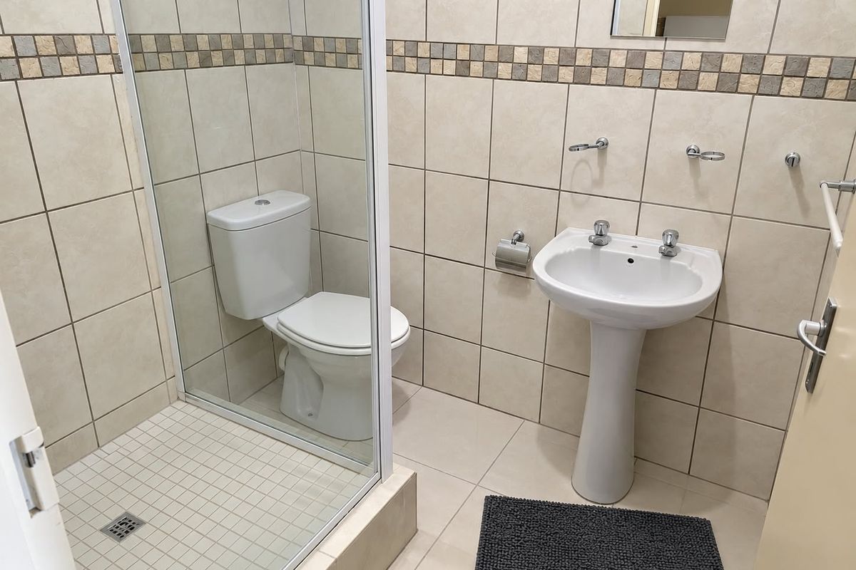 A functional bathroom featuring a pedestal basin, a glass-enclosed shower, and neutral wall tiling with a decorative mosaic border.