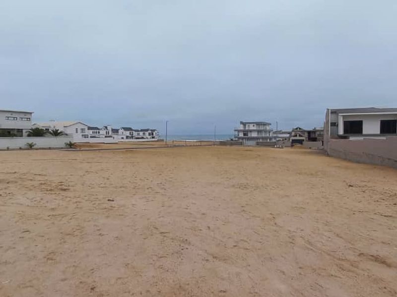 A vacant residential plot of land featuring sandy ground and boundary walls, situated in a coastal neighborhood with views toward the ocean.