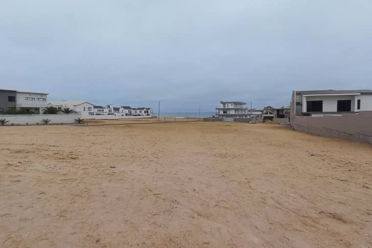 A vacant residential plot of land featuring sandy ground and boundary walls, situated in a coastal neighborhood with views toward the ocean.
