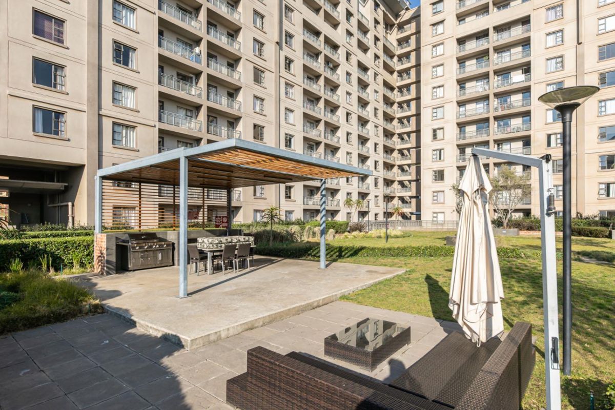 Communal outdoor entertainment area featuring a covered patio with a built-in braai, set within a landscaped garden of a large apartment complex.