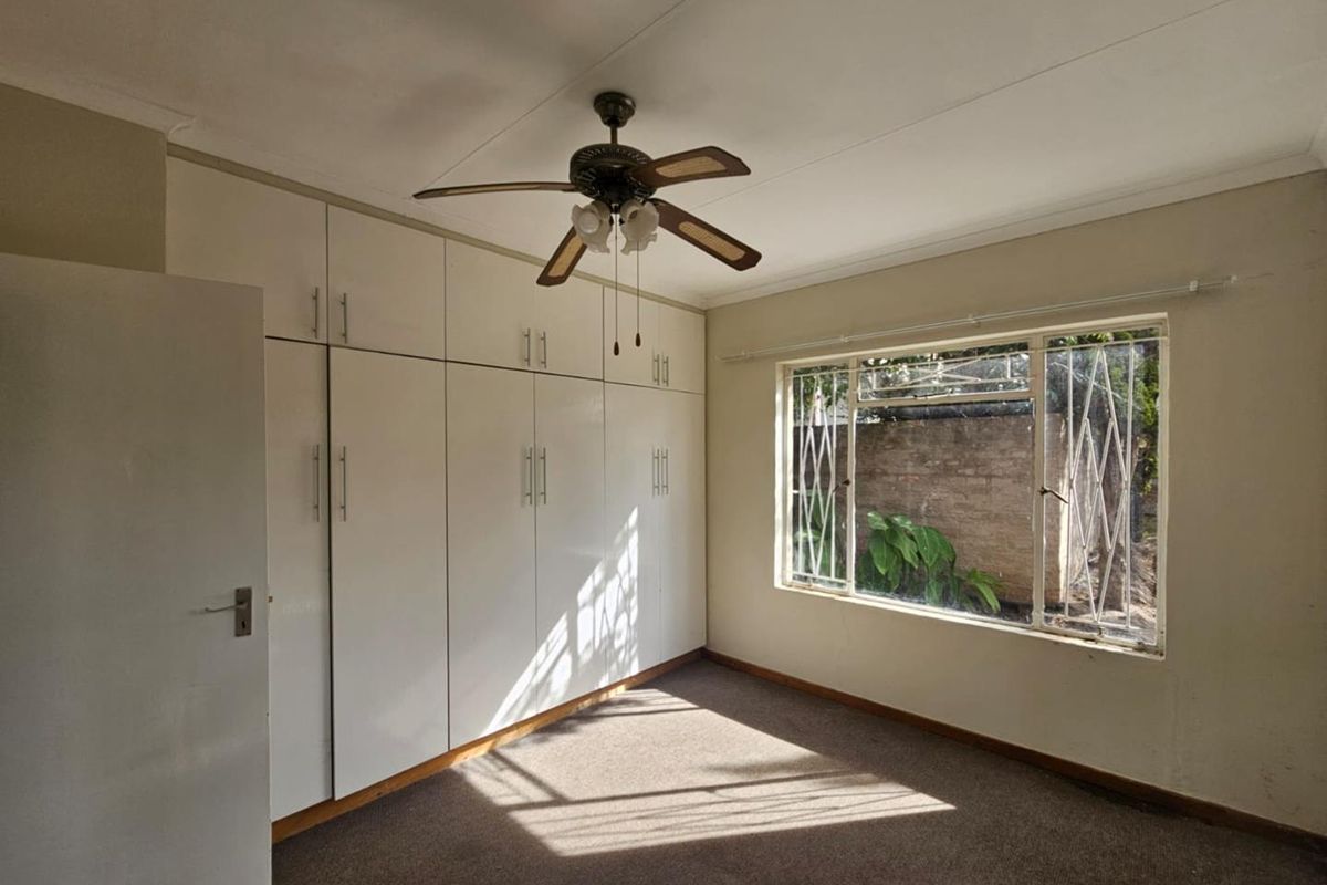 A bedroom featuring built-in white cabinetry, a ceiling fan, and a window with security bars overlooking the rear garden area.