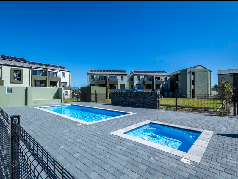 Communal outdoor area featuring a swimming pool, a smaller plunge pool, paved surround, and modern apartment buildings with solar panels in the background.