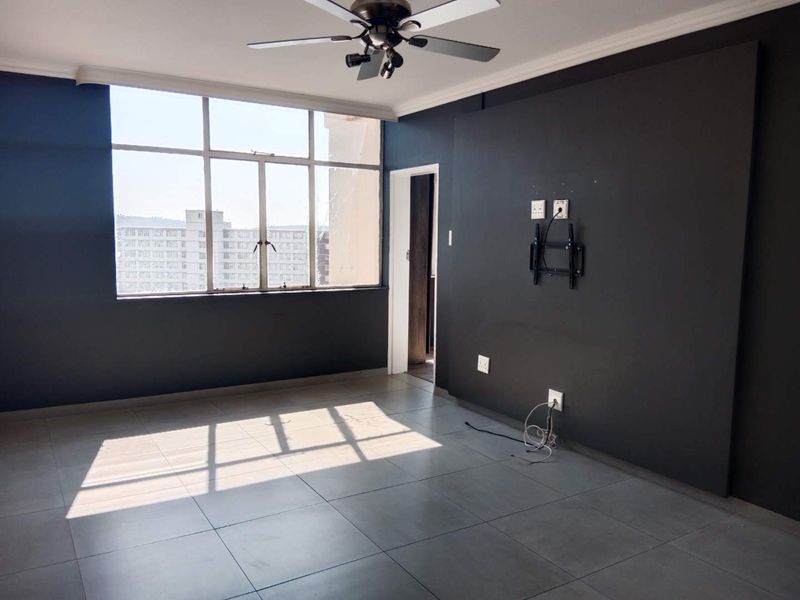 An empty living room featuring large windows, tiled flooring, a ceiling fan, and a dark accent wall with a mounted TV bracket.