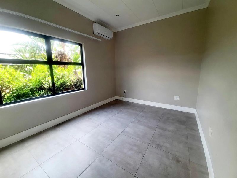 A clean, empty bedroom featuring large format grey floor tiles, a split-system air conditioner, and a large window overlooking the garden.