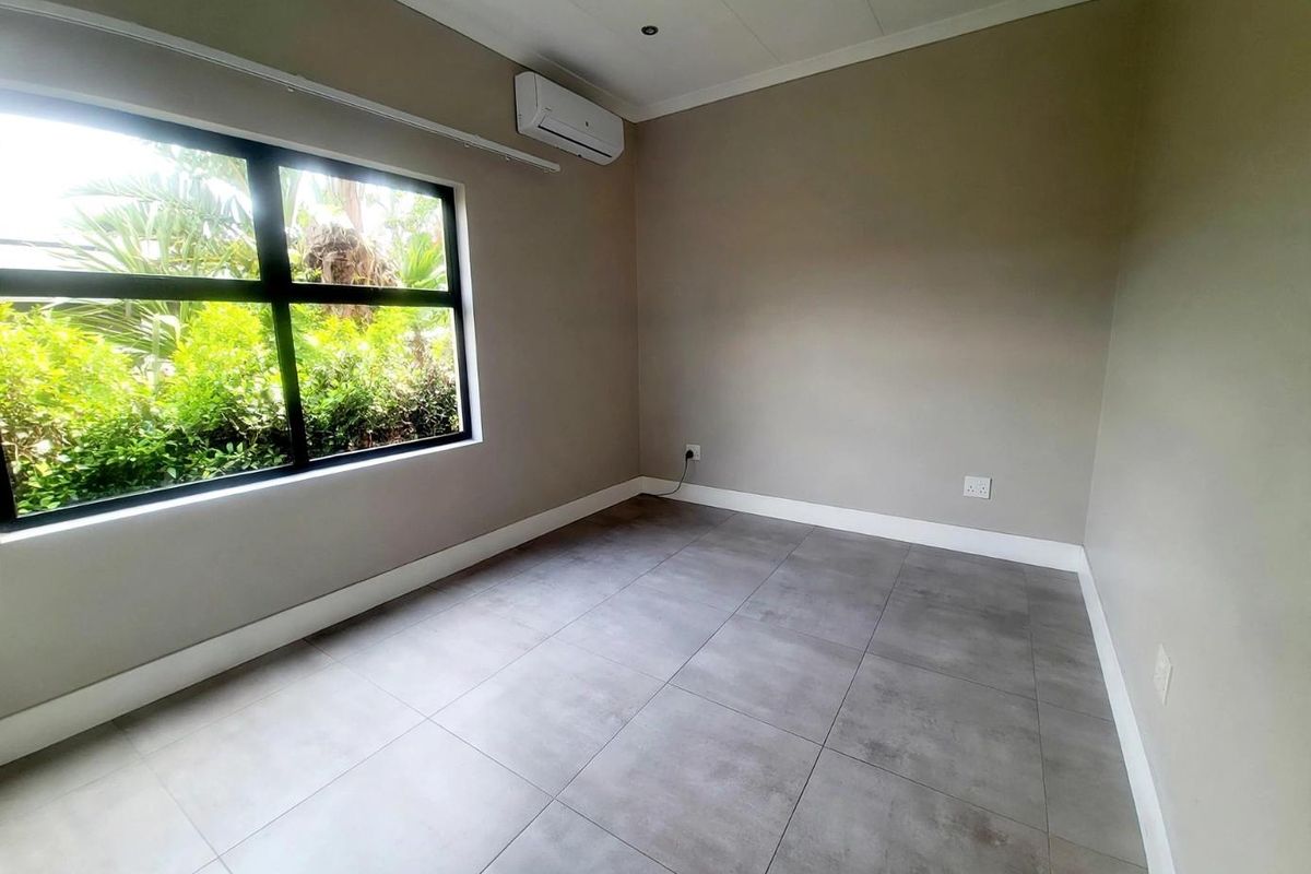 A clean, empty bedroom featuring large format grey floor tiles, a split-system air conditioner, and a large window overlooking the garden.