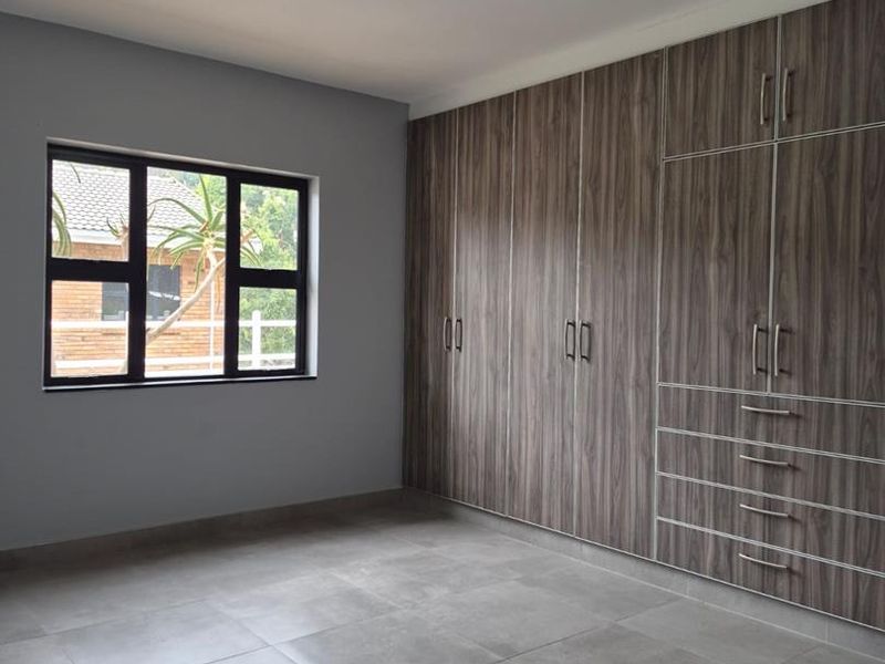 A clean, empty bedroom featuring modern grey floor tiles, a large built-in wooden wardrobe, and a dark-framed window.