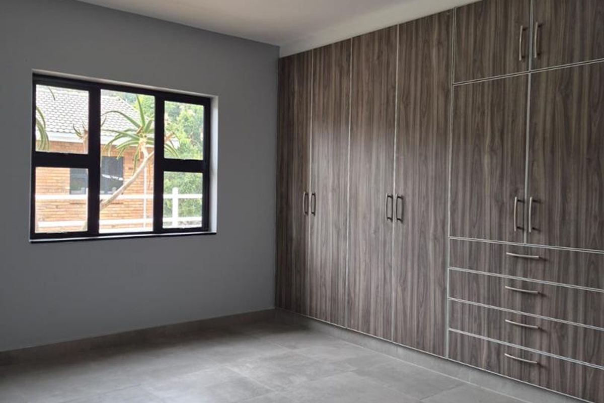 A clean, empty bedroom featuring modern grey floor tiles, a large built-in wooden wardrobe, and a dark-framed window.