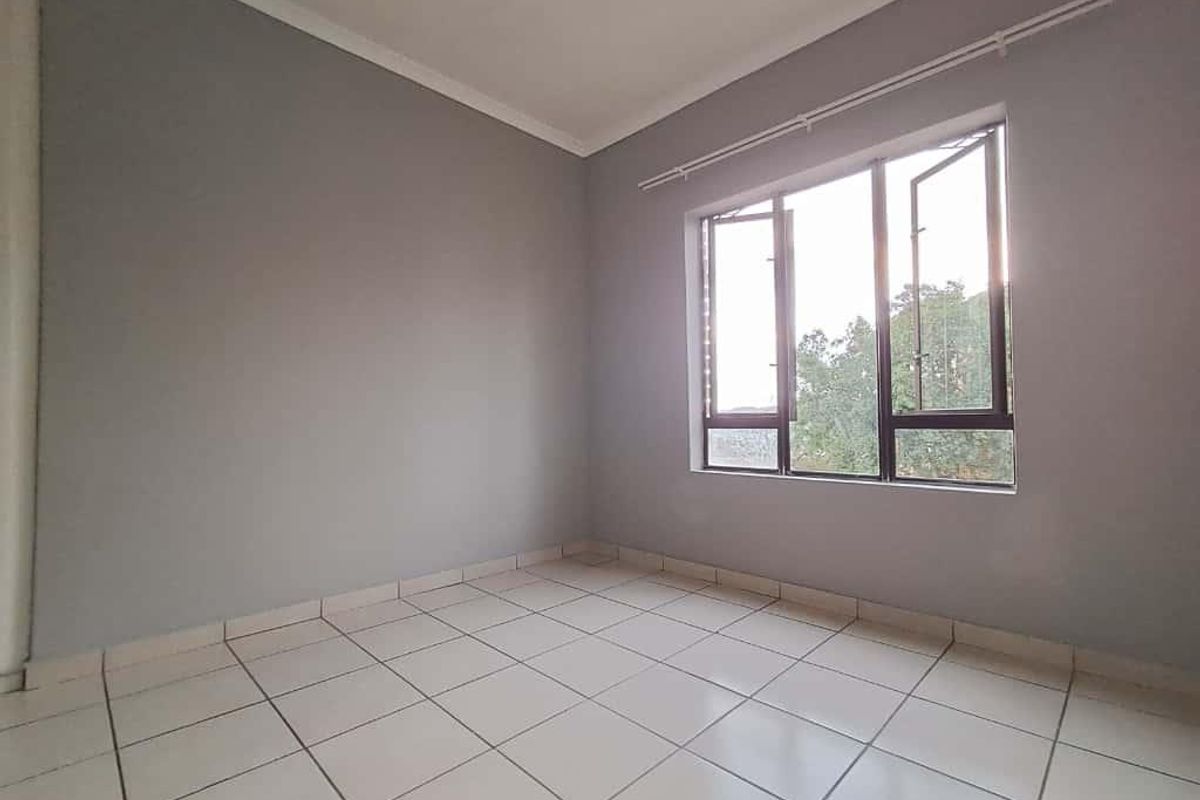 A bright, empty bedroom featuring neutral grey walls, tiled flooring, and a large window with metal frames.