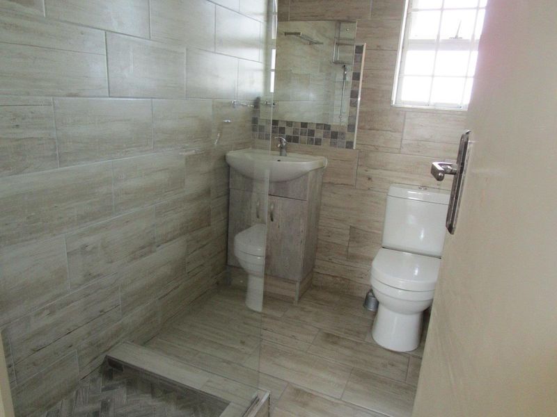 A modern bathroom featuring a walk-in shower with glass screen, vanity basin, and toilet, finished with neutral wood-look wall and floor tiles.