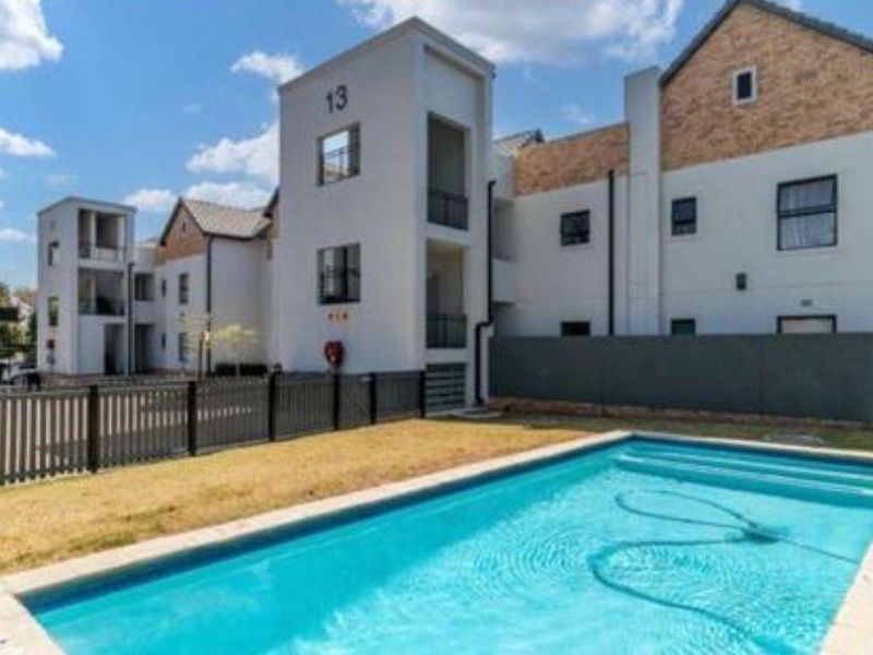 A well-maintained communal swimming pool area set against a modern multi-story apartment complex with secure perimeter fencing and covered parking.