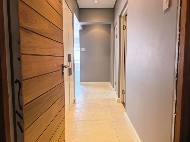 A clean, modern hallway featuring neutral grey walls, light-toned tiled flooring, and built-in storage cupboards, leading towards an open-plan living area.