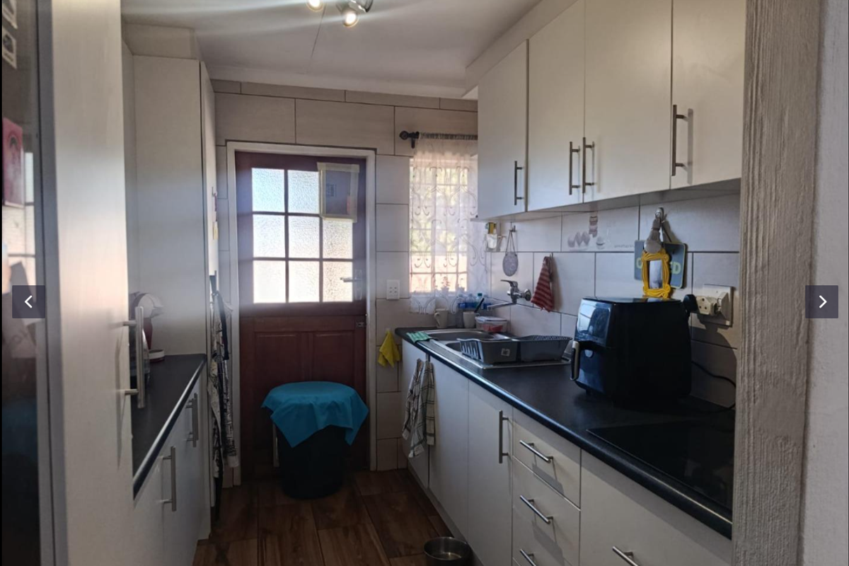A functional galley-style kitchen featuring white cabinetry, dark countertops, tiled backsplash, and wood-look flooring with direct access to the exterior.