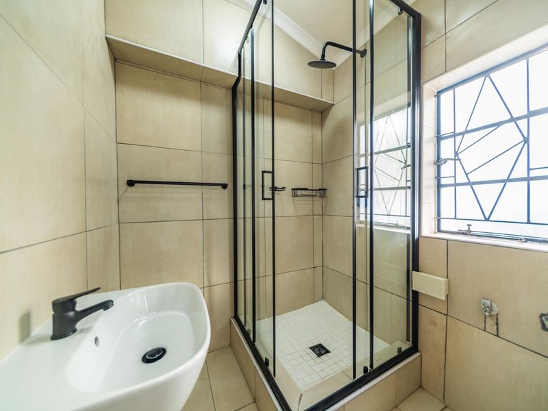 A modern bathroom featuring a glass-enclosed shower with a rain shower head, a contemporary basin with a black mixer tap, and neutral wall tiling.