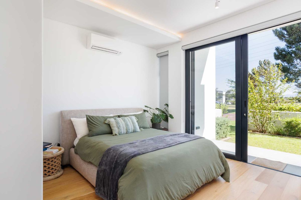 Modern bedroom featuring light timber flooring, recessed ceiling lighting, wall-mounted air conditioning unit, and large sliding glass doors opening to a private garden.