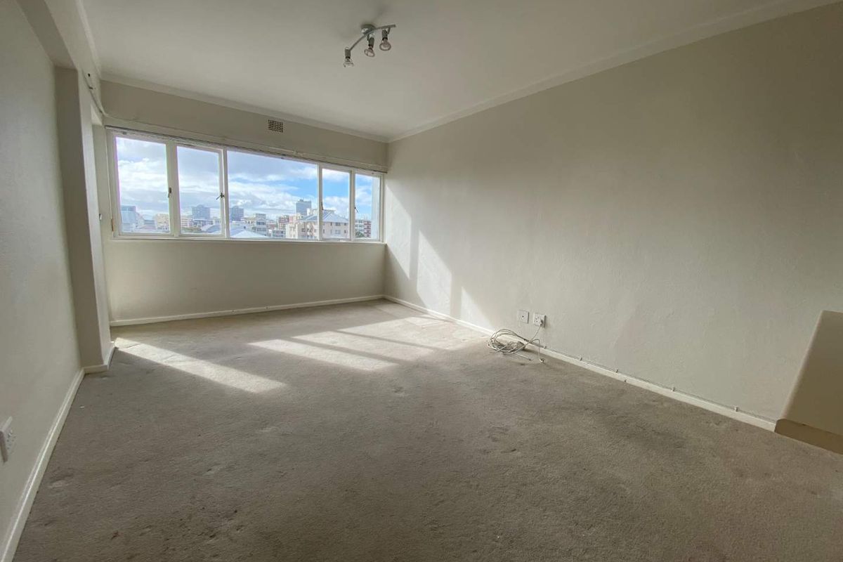 A spacious, sunlit bedroom featuring neutral walls, fitted carpeting, and large windows offering an urban city view.