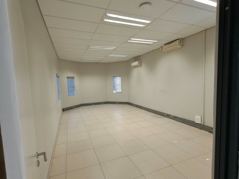 A spacious, empty office area featuring a suspended ceiling, recessed lighting, tiled floors, and wall-mounted air conditioning units.