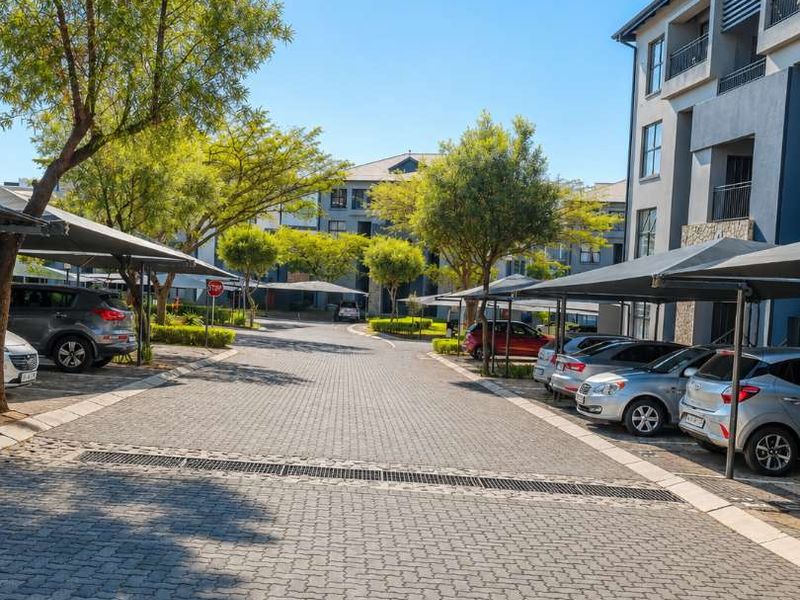 A well-maintained apartment complex exterior featuring paved driveways, covered parking bays, and established trees.