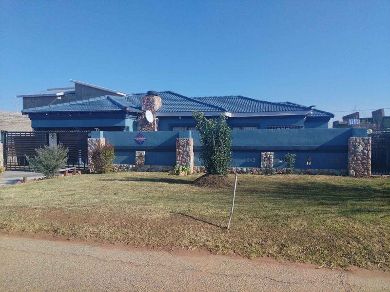 Front view of a residential house featuring a blue tiled roof, stone-clad boundary wall, and a secure metal driveway gate.