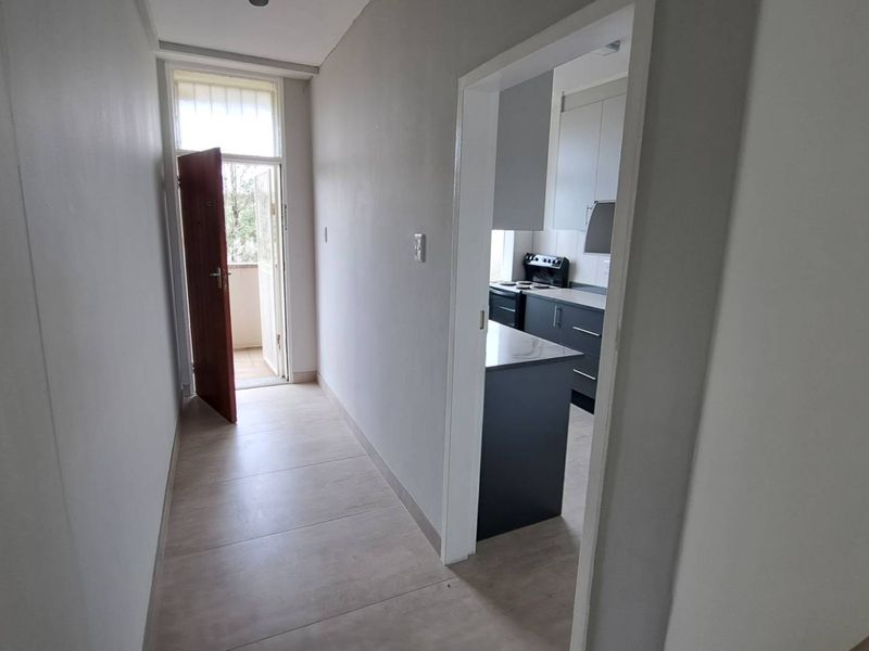 A bright hallway with neutral tiled flooring leading to an external door, with a view into a modern kitchen featuring dark cabinetry.