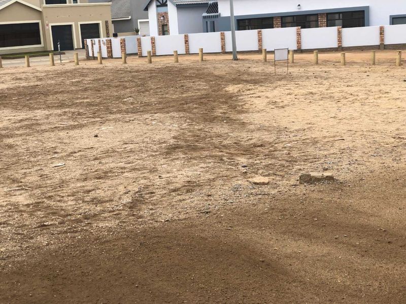 A vacant residential plot featuring a level sandy surface, enclosed by wooden bollards and a boundary wall, situated in a developed residential area.
