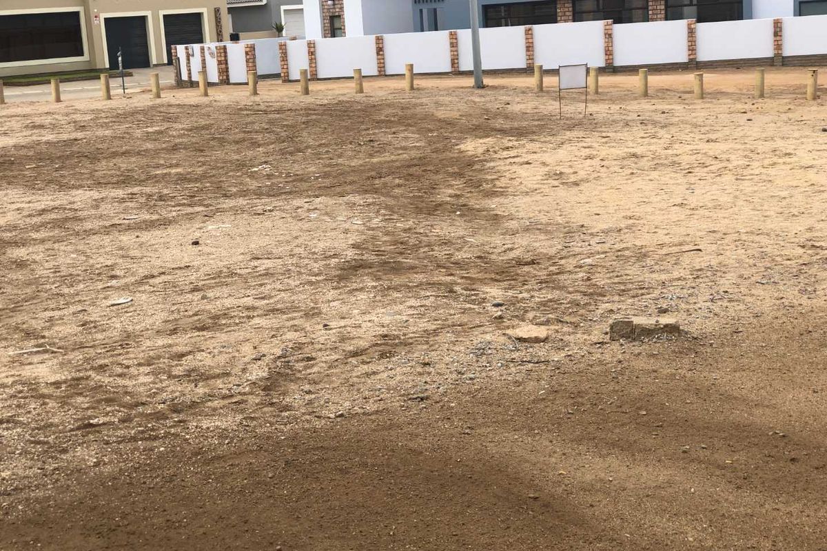 A vacant residential plot featuring a level sandy surface, enclosed by wooden bollards and a boundary wall, situated in a developed residential area.