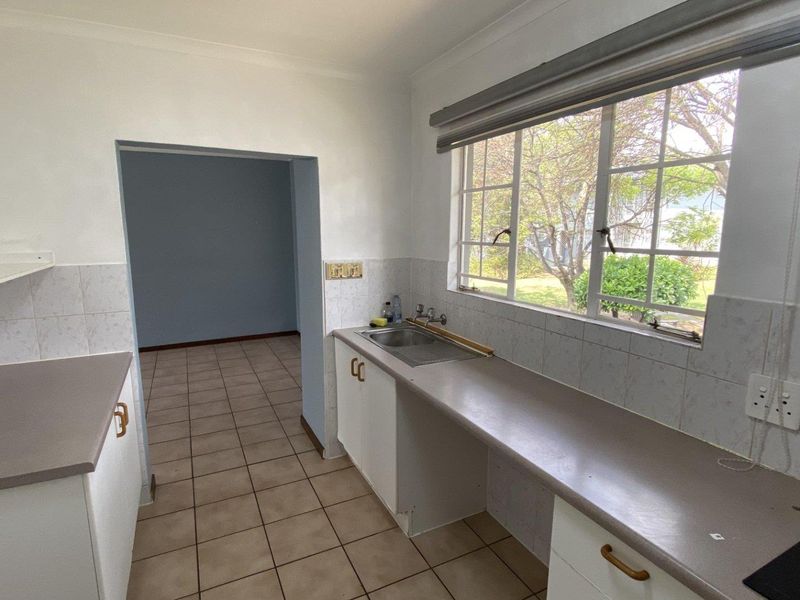 A functional, empty kitchen featuring laminate countertops, a stainless steel sink, tiled backsplash, and ceramic floor tiles with a view of the garden.