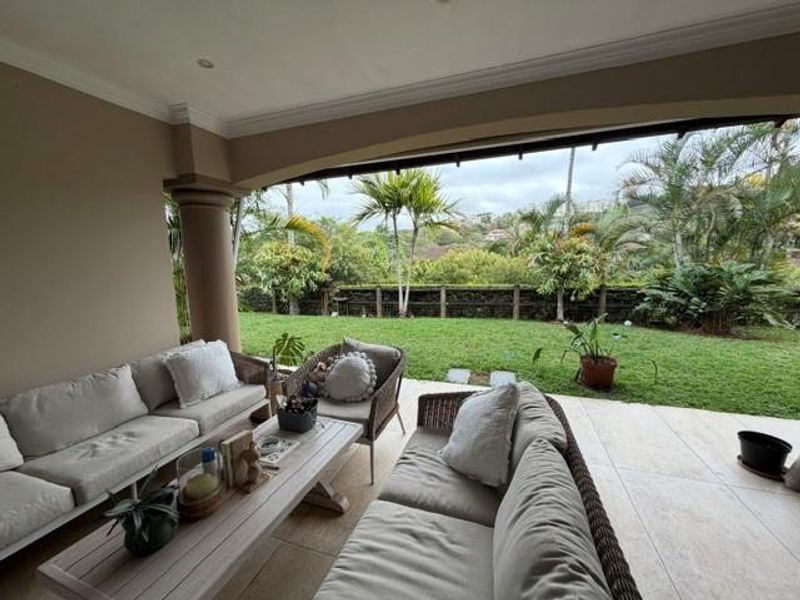 A spacious covered patio featuring tiled flooring and recessed lighting, overlooking a private garden with lush greenery and a scenic landscape view.
