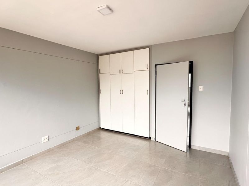 A clean, empty bedroom featuring neutral grey walls, tiled flooring, and white built-in cupboards with overhead storage.