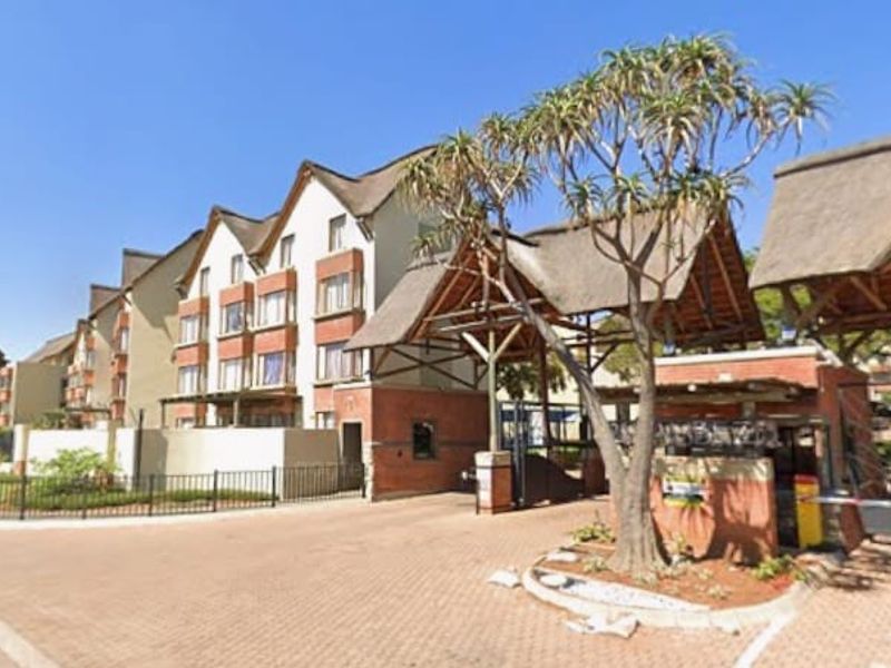 Exterior view of the apartment complex entrance featuring a brick-paved driveway, security gatehouse with thatched roofing, and perimeter fencing.