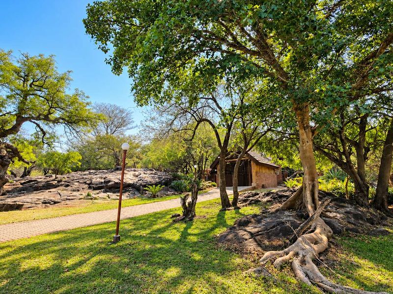 A beautifully landscaped garden featuring mature trees, natural rock formations, and a paved walkway leading towards a rustic structure.