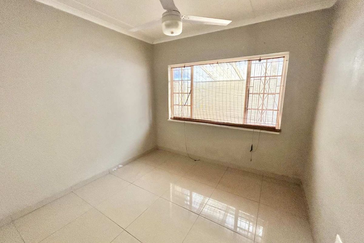 A bright, empty bedroom featuring neutral tiled flooring, a ceiling fan, and a window fitted with security bars.