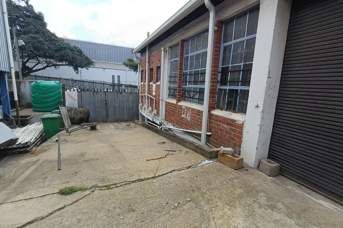 Exterior side view of an industrial property featuring brick facade, steel-framed windows with security bars, and a concrete yard area.