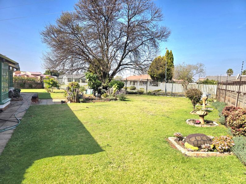 A spacious backyard featuring a well-maintained lawn, mature trees, and established garden beds enclosed by secure metal perimeter fencing.