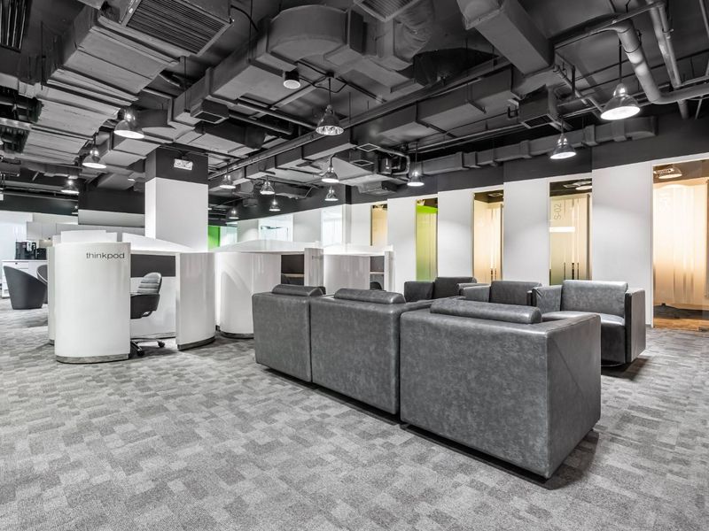 Modern open-plan office space featuring exposed industrial ceiling ductwork, carpeted flooring, and integrated private work pods with glass partition doors.
