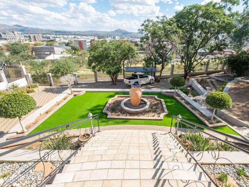 A well-maintained terraced garden featuring an artificial lawn, central water feature, stone staircase with wrought iron railings, and panoramic city views.