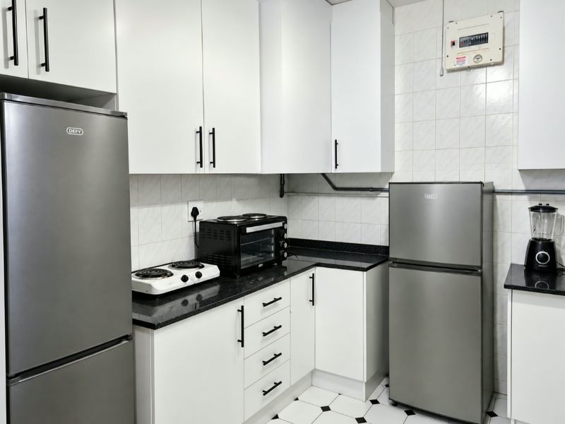 A compact kitchen featuring white cabinetry, black granite countertops, tiled backsplash, and tiled flooring, with an accessible electrical distribution board.