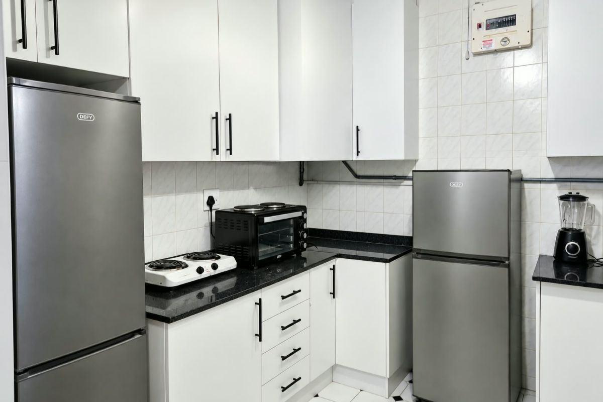 A compact kitchen featuring white cabinetry, black granite countertops, tiled backsplash, and tiled flooring, with an accessible electrical distribution board.