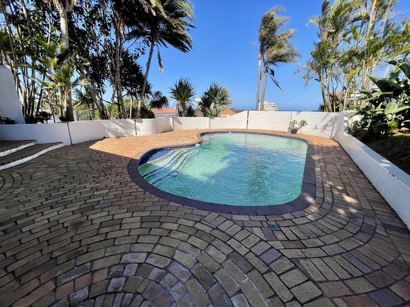 Outdoor pool area featuring a curved swimming pool surrounded by brick paving and a low retaining wall, set within a landscaped garden.