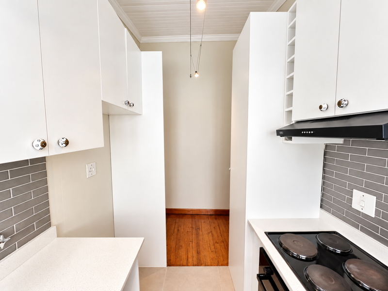 A clean, modern kitchen featuring white cabinetry, grey subway tile backsplash, solid surface countertops, and integrated appliances, leading to a timber-floored area.