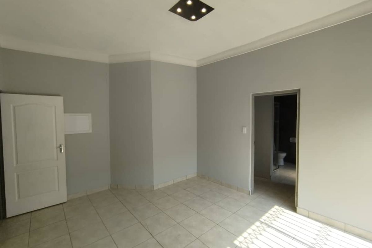 A bright, empty bedroom featuring neutral grey walls, tiled flooring, a modern flush-mount ceiling light, and an adjoining en-suite bathroom.