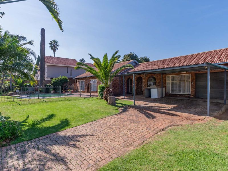 Rear view of the property featuring a brick facade, covered patio area, paved driveway, and a swimming pool set within a landscaped garden.