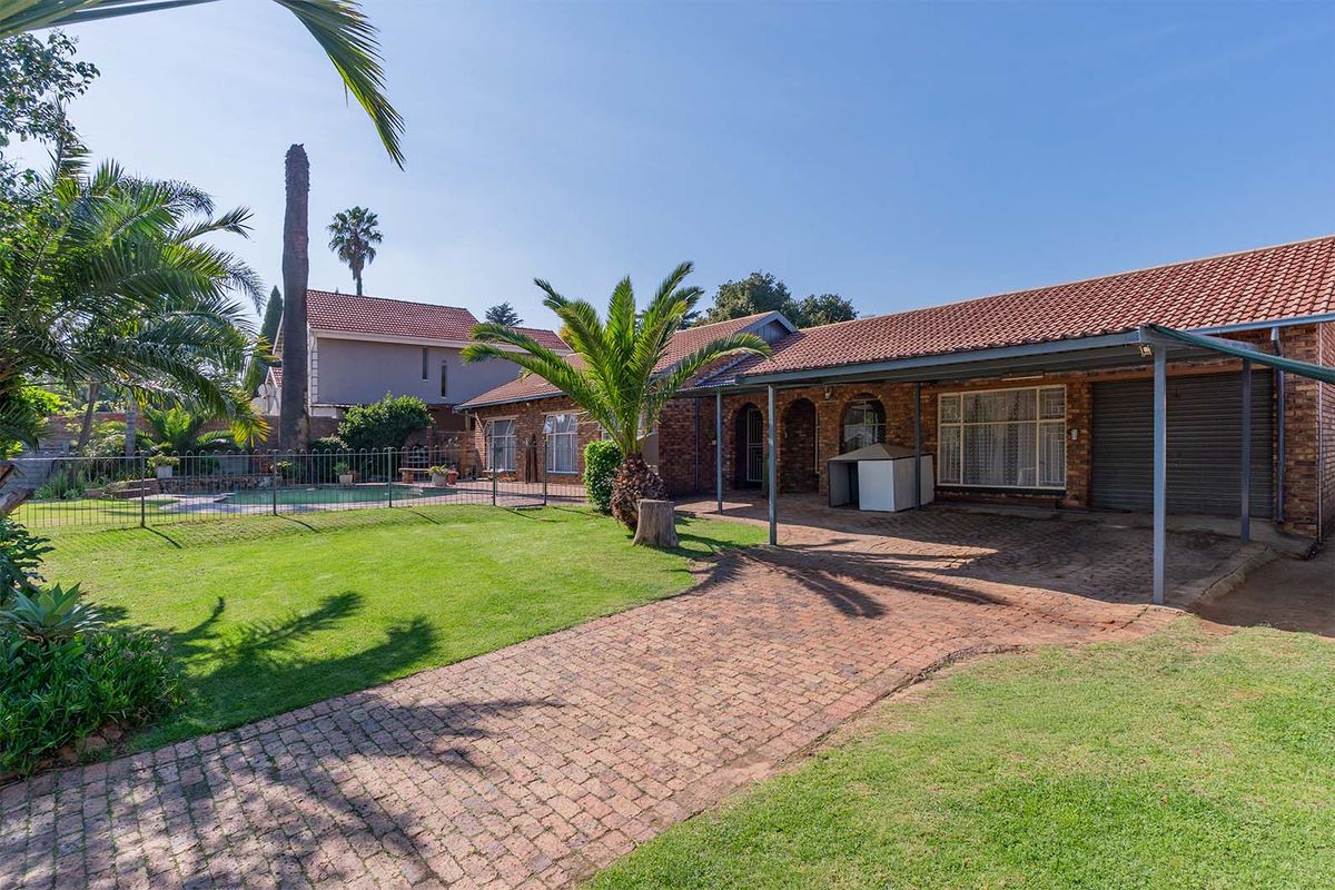 Rear view of the property featuring a brick facade, covered patio area, paved driveway, and a swimming pool set within a landscaped garden.