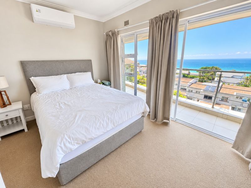 A well-maintained bedroom featuring carpeted flooring, a wall-mounted air conditioner, and large sliding glass doors opening onto a balcony with ocean views.