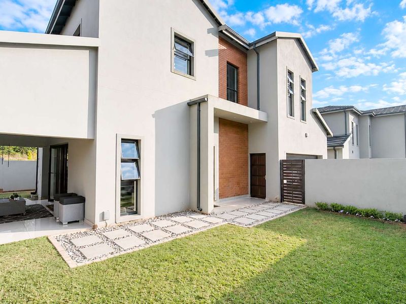 Rear view of a modern cluster home featuring a covered patio, paved walkway, landscaped garden, and a combination of plaster and brick exterior finishes.