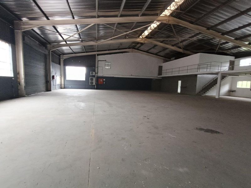 Spacious industrial warehouse featuring high ceilings with exposed steel trusses, concrete flooring, a roller shutter door, and an integrated mezzanine level.