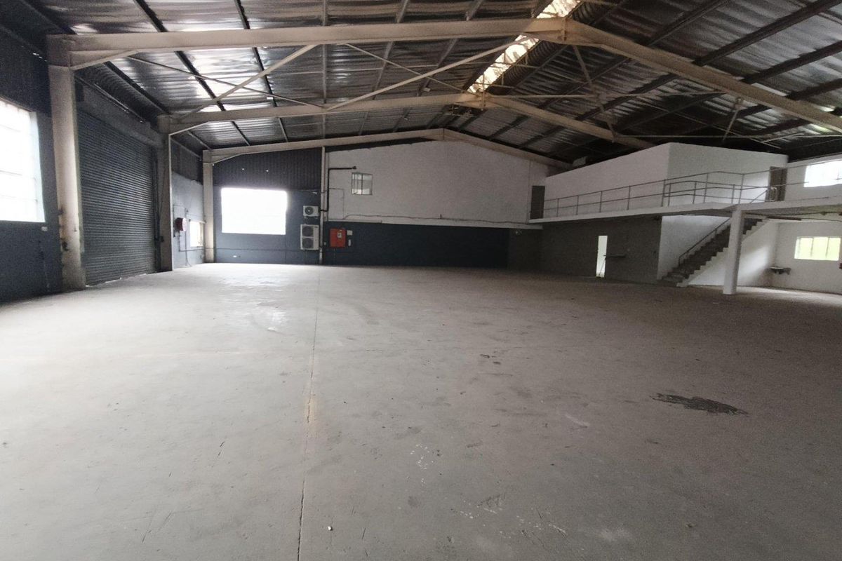 Spacious industrial warehouse featuring high ceilings with exposed steel trusses, concrete flooring, a roller shutter door, and an integrated mezzanine level.