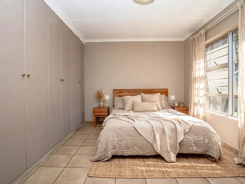 A well-maintained bedroom featuring neutral tones, extensive built-in cupboard storage, tiled flooring, and natural light from a large window.